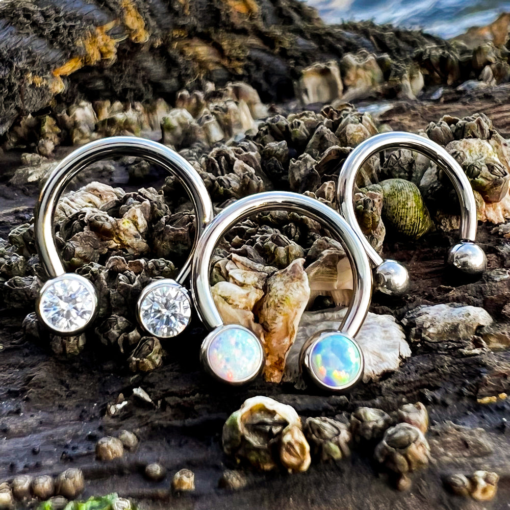 Three threadless titanium circular barbells with cubic zirconia faceted gem ends, white opal cabochon gem ends, and titanium ball ends.